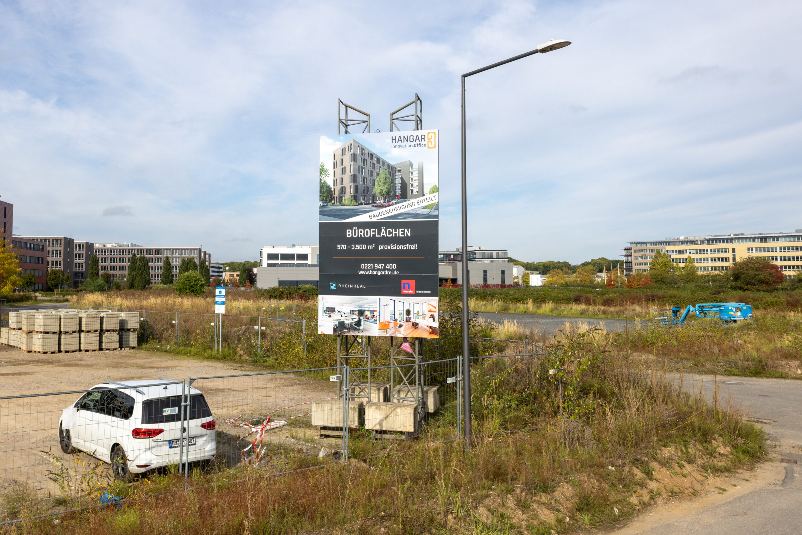 "Großes Bauschild für ""Hangar 3"" in Köln mit Informationen zu Büroflächen, umgeben von einer Baustelle und einem weißen Auto."
