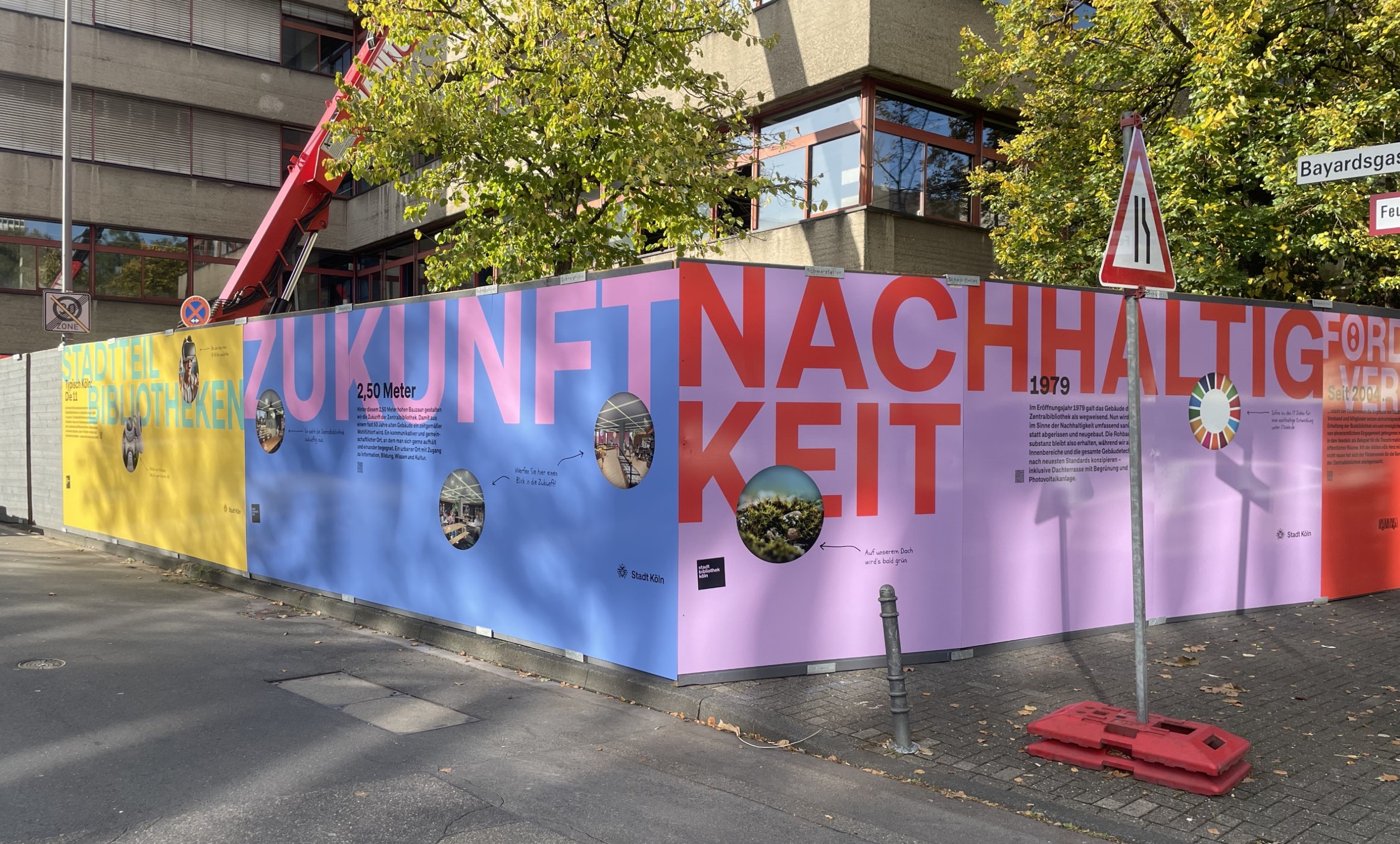 "Bunter Bauzaun mit den Worten ""ZUKUNFT NACHHALTIGKEIT"" und Informationen zu Stadtteilbibliotheken, zeigt nachhaltiges Baustellenmarketing."
