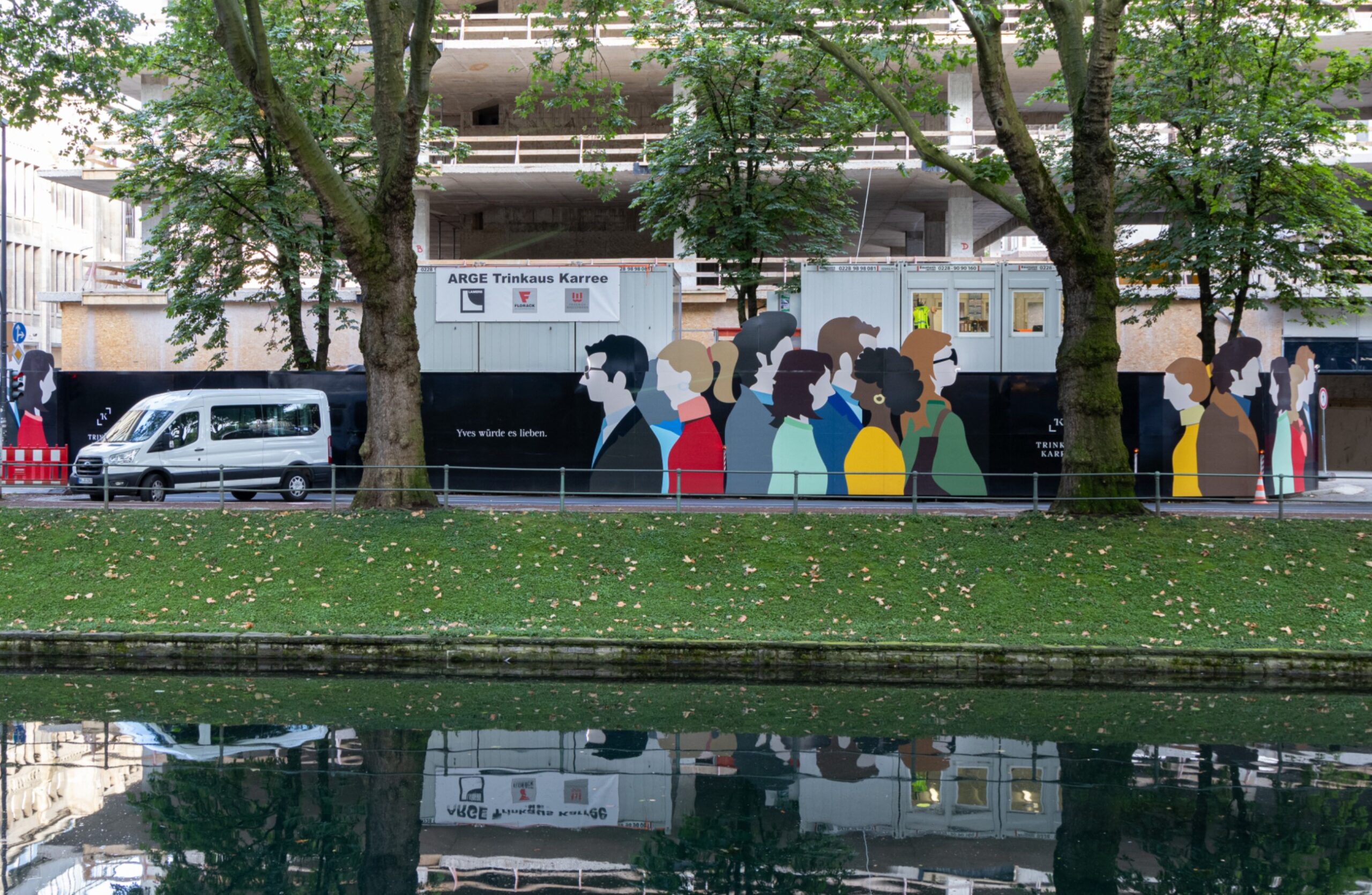 "Langer Bauzaun mit farbigen Silhouetten von Menschen und dem Slogan ""Vom Wandel des Lebens"", spiegelt sich im Wasser."
